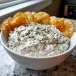 A white bowl filled with creamy dill dip, garnished with fresh dill, served with ruffled potato chips.