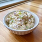 A close-up view of a creamy ham salad recipe mixed with celery chunks, served in a small white bowl on a wooden surface.