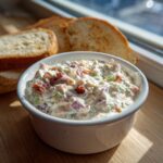 Close-up of a white bowl filled with creamy hoagie dip, featuring visible chunks of meat and vegetables, served next to toasted bread.