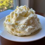 A large mound of fluffy, pale yellow creamy mashed potatoes served on a white plate near a window.