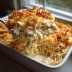 A spoonful of creamy monterey chicken spaghetti being lifted from a white baking dish, topped with melted cheese and crispy onions.