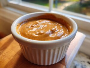 Close-up of thick, creamy peanut sauce in a white fluted ramekin, set near a bright window.