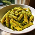 Close-up of penne pasta coated generously in vibrant green creamy pesto pasta sauce in a white bowl.