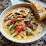 A bowl of creamy philly cheese steak soup topped with strips of steak, red and green peppers, served with a piece of crusty bread.