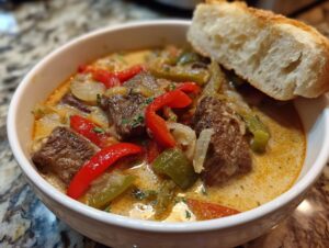 A close-up of a bowl of creamy philly cheese steak soup filled with chunks of beef, red peppers, green peppers, and onions, topped with a piece of crusty bread.
