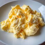Close-up of perfectly creamy and soft scrambled eggs piled high on a simple white plate.