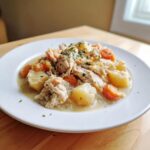 A white plate holding a serving of creamy slow cooker chicken stew with shredded chicken, potatoes, and carrots, garnished with parsley.
