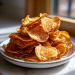 A tall stack of golden, crispy homemade potato chips seasoned with spices, served on a small white plate.