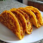 A row of four golden, crispy potato tacos filled with mashed potato mixture, served on a white plate.