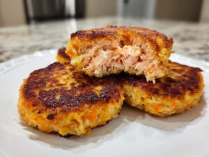 Close-up of three golden-brown, crispy salmon cakes on a white plate, one cut open to show the flaky salmon interior.