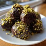 A pile of rich, dark Dubai chocolate balls dipped in ganache and generously sprinkled with chopped green pistachios.