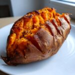 A close-up of a perfectly baked sweet potato, split open to reveal fluffy orange flesh seasoned with dark spices.