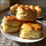 Three tall, flaky buttermilk biscuits stacked on a white plate, showing their layered texture and golden tops.
