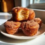 A stack of golden brown french toast muffins coated generously in cinnamon sugar, with one muffin broken open to show the fluffy interior.