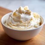A close-up of light, fluffy vanilla buttercream frosting swirled high in a white bowl on a wooden surface.