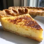 Close-up of a slice of silky custard pie with a golden, flaky crust and a cinnamon-dusted top.