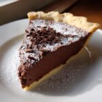 A decadent slice of french silk pie with a thick chocolate filling, topped with powdered sugar and chocolate shavings.