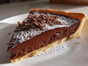 A close-up of a slice of rich french silk pie featuring a thick chocolate filling, powdered sugar, and chocolate shavings.