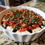 A white scalloped dish filled with creamy bruschetta dip, topped generously with diced tomatoes, fresh basil, and a balsamic glaze.