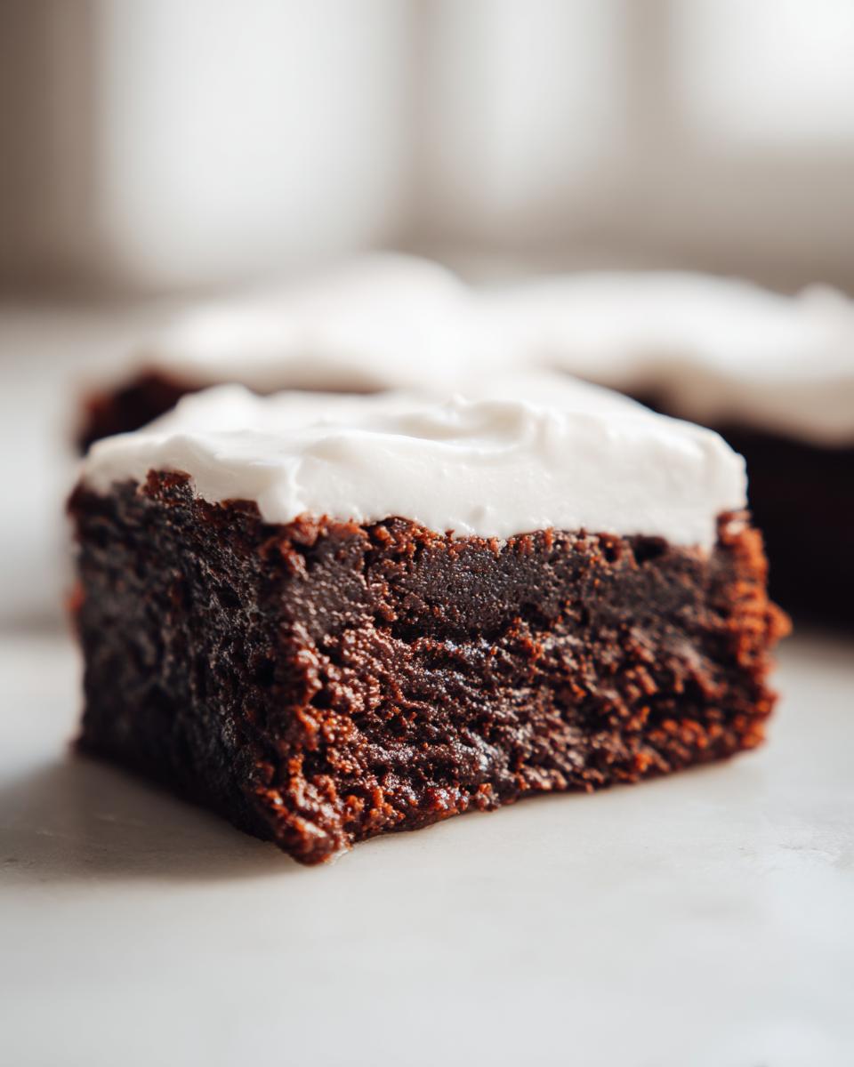 A close-up shot of a rich, dark square of gingerbread brownies topped with thick white frosting.