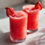 Two tall glasses filled with bright red, frozen strawberry daiquiri, each garnished with a fresh strawberry slice.