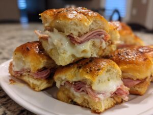 A stack of freshly baked, cheesy grinder sliders featuring melted white cheese and ham on glossy buns.