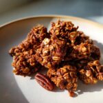A pile of rich, dark brown gingerbread granola clusters on a light plate, next to a single pecan.