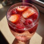 Close-up of a glass filled with bubbling pink champagne punch, garnished with fresh strawberries and raspberries.
