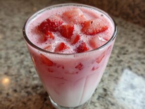 Close-up of a tall glass filled with homemade strawberry milk, topped with frothy foam and chunks of fresh strawberries.