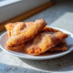 Close-up of several golden brown buñuelos generously coated in cinnamon sugar, resting on a white plate.