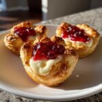 Three golden, flaky puff pastry brie bites topped with melted brie and bright red jam, sitting on a white plate.