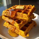 Close-up of a stack of three fluffy pumpkin waffles being drizzled with syrup and topped with butter.