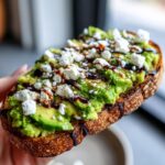A close-up of gourmet avocado toast topped with crumbled feta cheese, spices, and a balsamic glaze drizzle.
