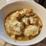 Close-up of a white bowl filled with rich gravy, tender chicken pieces, and fluffy dumplings in a classic chicken and dumpling dish.
