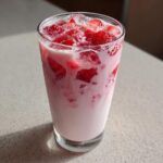 A tall glass filled with pink strawberry milk and chunks of fresh strawberries and ice.