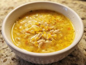 A close-up view of a white bowl filled with bright yellow Italian penicillin soup, featuring shredded chicken and orzo pasta.