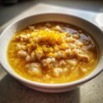 A close-up of a bowl of vibrant yellow Italian penicillin soup, featuring small pasta shapes and topped with bright lemon zest.