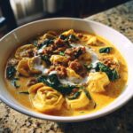 A close-up of a rich, creamy bowl of Italian sausage tortellini soup with spinach and dollops of cream.