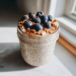 A glass jar filled with creamy chia seed pudding, topped with fresh blueberries, cinnamon, and nut pieces.