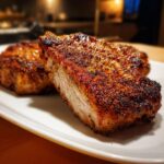 Close-up of two thick, juicy air fryer pork chops with a heavily seasoned, dark brown crust served on a white plate.