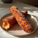Two crispy, sugar-coated korean corn dogs on a plate, one showing a dramatic, melted cheese pull.