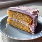 A close-up of a moist slice of lavender cake with light purple lavender frosting between layers and on top.
