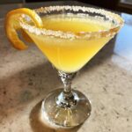 A bright yellow lemon drop martini in a stemmed glass with a sugar-rim and orange twist garnish.