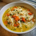 Close-up of a white bowl filled with Lemon Ginger Turmeric Chicken and Rice Soup, showing shredded chicken, rice, and bright orange carrots.
