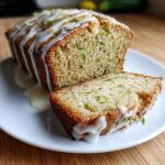 A loaf of moist lemon zucchini bread, drizzled with white glaze and topped with zest, with one slice cut and resting beside it.