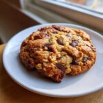 A thick, golden-brown levain bakery chocolate chip cookies studded with dark chocolate chunks and walnuts, resting on a white plate.