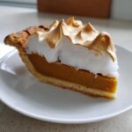 A close-up of a thick slice of butterscotch pie with a rich orange filling and toasted meringue topping on a white plate.