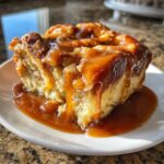 A close-up of a moist slice of apple bread pudding generously drizzled with thick caramel sauce, sitting on a white plate.