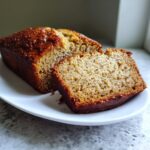 Close-up of a freshly baked loaf of banana bread recipe, with one thick slice cut and resting against the main loaf on a white plate.