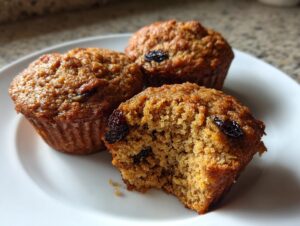 Close-up of three moist bran muffins on a white plate, one is broken open showing the texture and raisins.